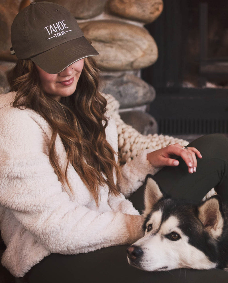 Woman wearing a cap with 'TANOE' logo sitting next to a dog in front of a stone wall.